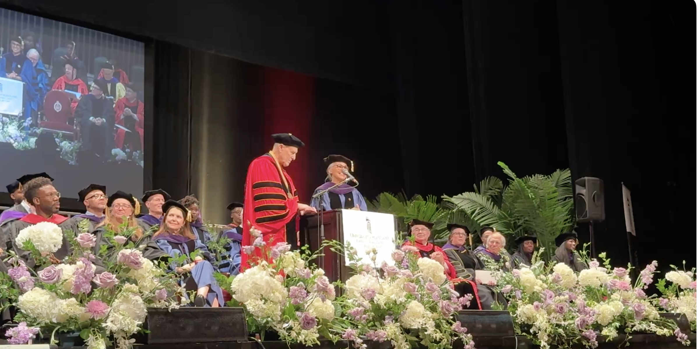 Two speakers stand at the podium on a stage wearing academic regalia with faculty seated behind them