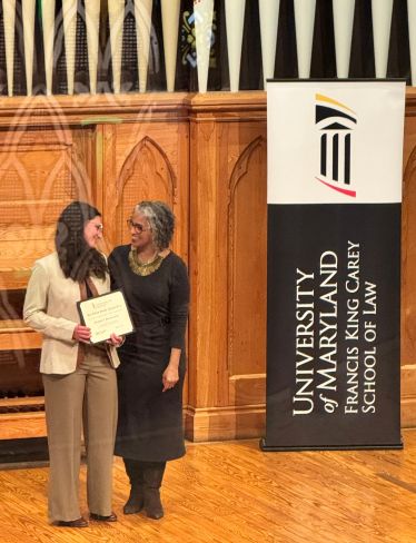 Award recipient stands on a stage holding a certificate next to the dean