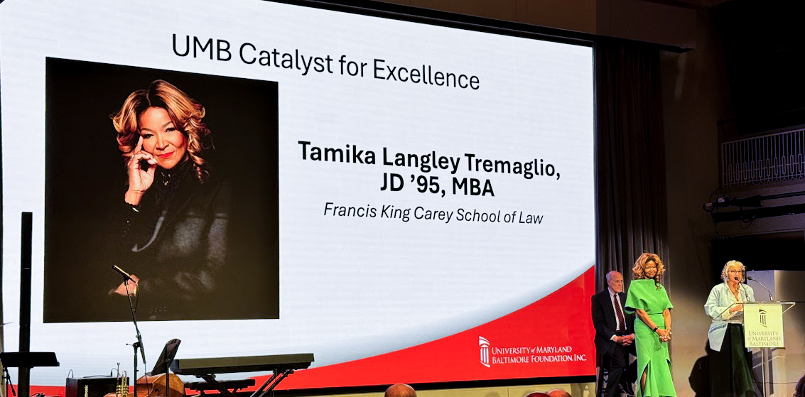 Three people dressed formally on a stage in front of a screen announcing awardee